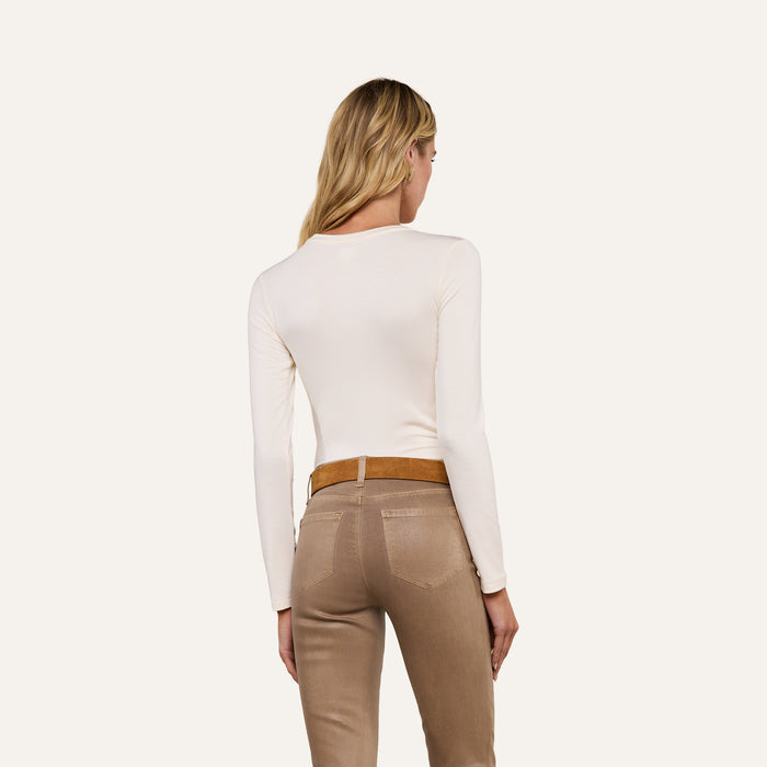 A person with long blonde hair is shown from the back, wearing the Tess Long Sleeve Tee in a slim fit cream shade and beige pants with a brown belt, standing against a plain light background.