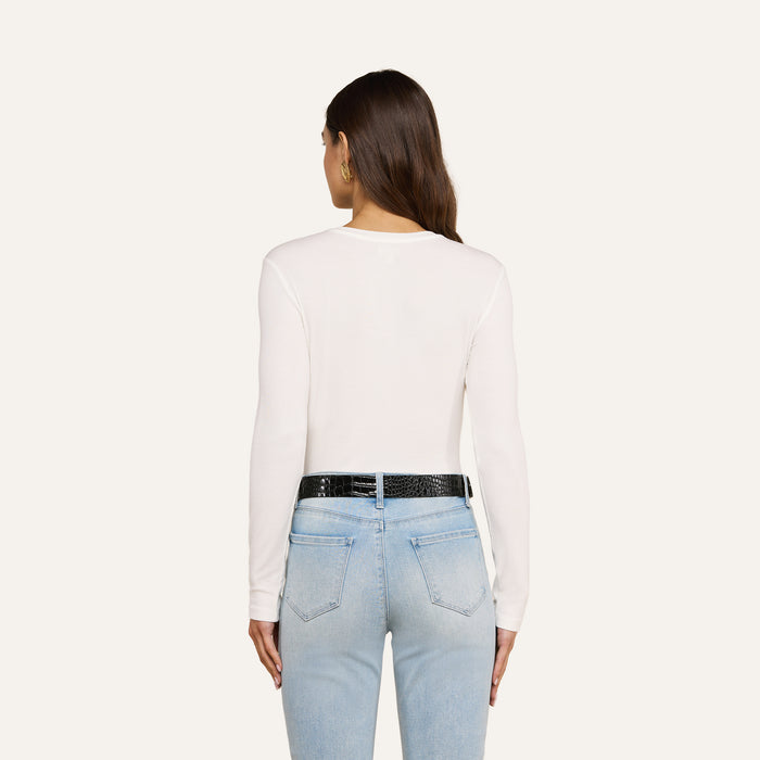 A woman with long brown hair is shown from the back, wearing the Tess Long Sleeve Tee in white, paired with light blue jeans and a black belt, standing against a plain light background.
