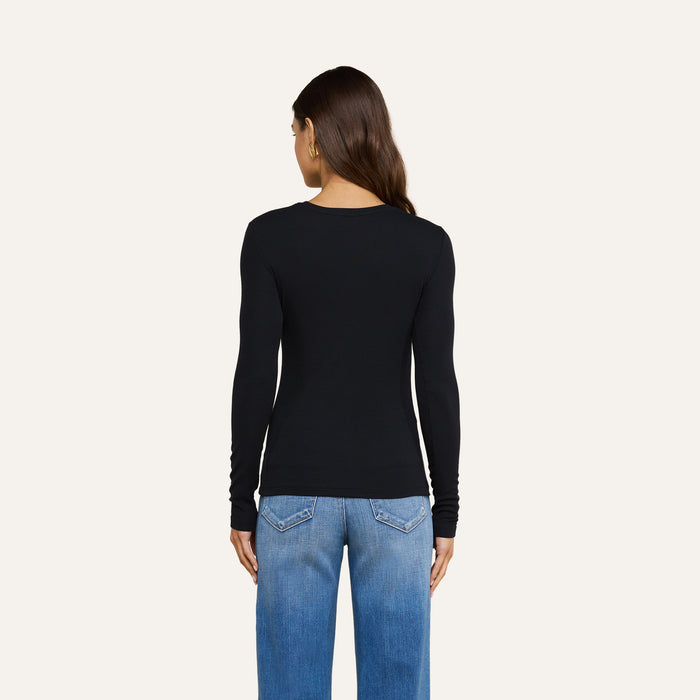 A woman with long brown hair, dressed in the Tess Long Sleeve Tee and blue jeans, stands facing away from the camera against a plain light background.