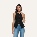 A woman wearing the Tessa Vest, a sleeveless black top with gold buttons, paired with blue jeans stands against a plain light background, touching her hair.