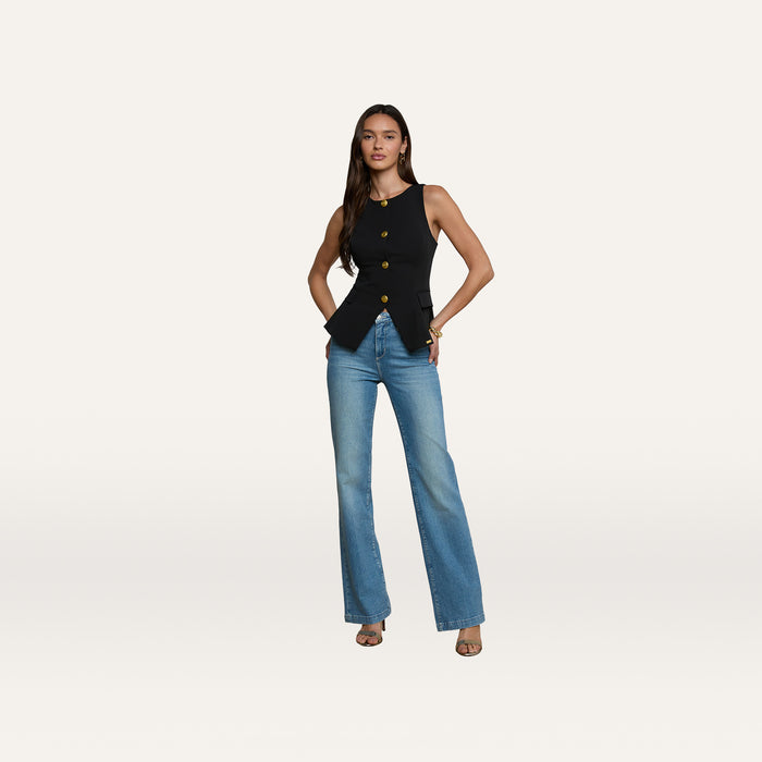 A woman stands with her hands in her pockets, wearing the Tessa Vest—a sleeveless black top with gold buttons—paired with blue flared jeans crafted from stretch suit fabric and open-toe heels, set against a plain white background.