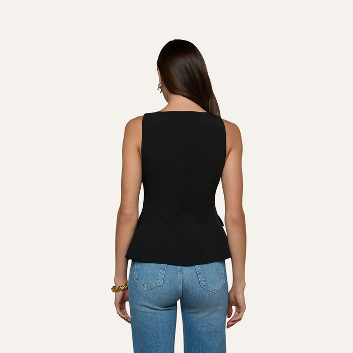A woman with long brown hair is shown from the back, wearing the Tessa Vest—a sleeveless suit top with gold buttons—paired with blue jeans against a plain light background.