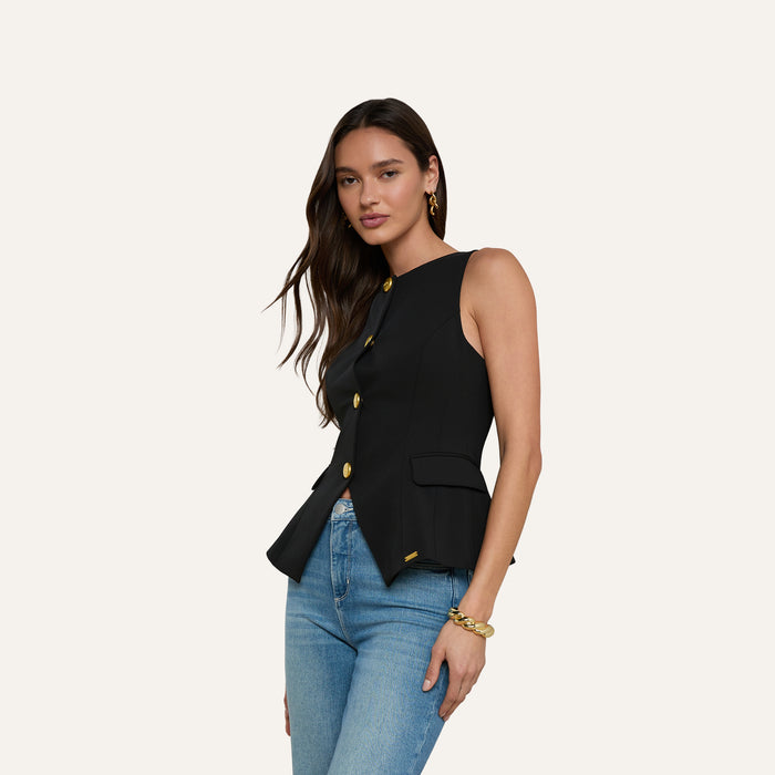 A woman with long brown hair models the Tessa Vest—a sleeveless, suit-style black vest crafted from stretch suit fabric and featuring gold buttons—paired with blue jeans and gold jewelry, standing against a plain light background.