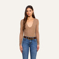 A woman with long brown hair is standing against a plain light background, wearing the Thalassa High-Leg Bodysuit featuring sexy mesh and sheer nude long sleeves, paired with blue jeans, a wide brown belt with large gold buckles, and gold hoop earrings.
