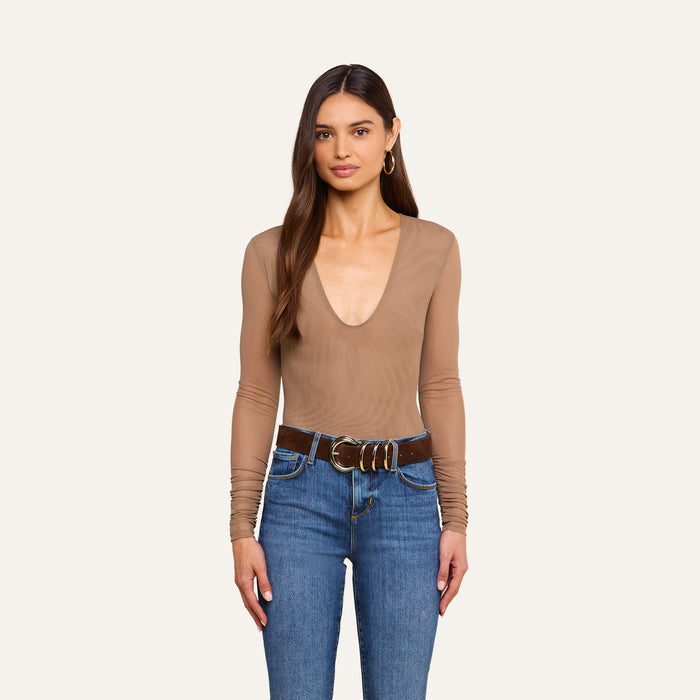 A woman with long brown hair is standing against a plain light background, wearing the Thalassa High-Leg Bodysuit featuring sexy mesh and sheer nude long sleeves, paired with blue jeans, a wide brown belt with large gold buckles, and gold hoop earrings.