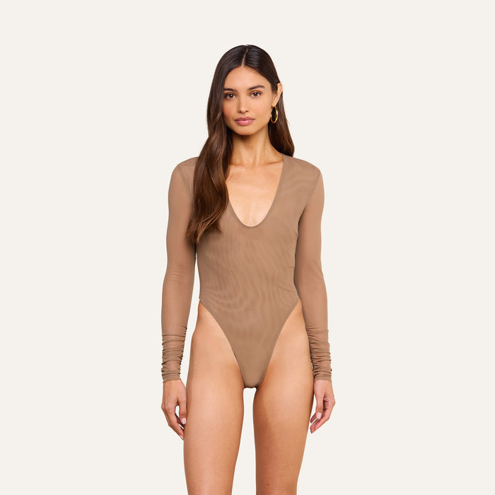 A woman models the Thalassa High-Leg Bodysuit, featuring sheer mesh fabric, long sleeves, and a deep scoop neckline. She stands against a plain light background, her long brown hair cascading over her shoulders as she looks at the camera.