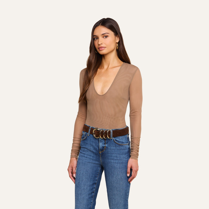 A woman with long brown hair wears the Thalassa High-Leg Bodysuit paired with blue jeans and a brown belt, standing against a plain light background.