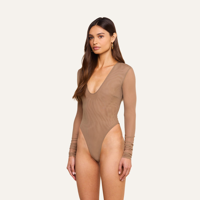 A woman stands against a plain background wearing the Thalassa High-Leg Bodysuit—a fitted, long-sleeve mesh piece with a deep neckline in sheer beige. She has long brown hair and a neutral expression, embodying effortless French style.