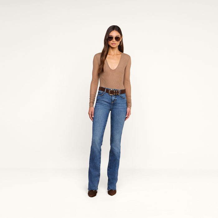 A woman stands against a plain background, wearing sunglasses, blue jeans with a brown belt, brown shoes, and the Thalassa High-Leg Bodysuit in tan.