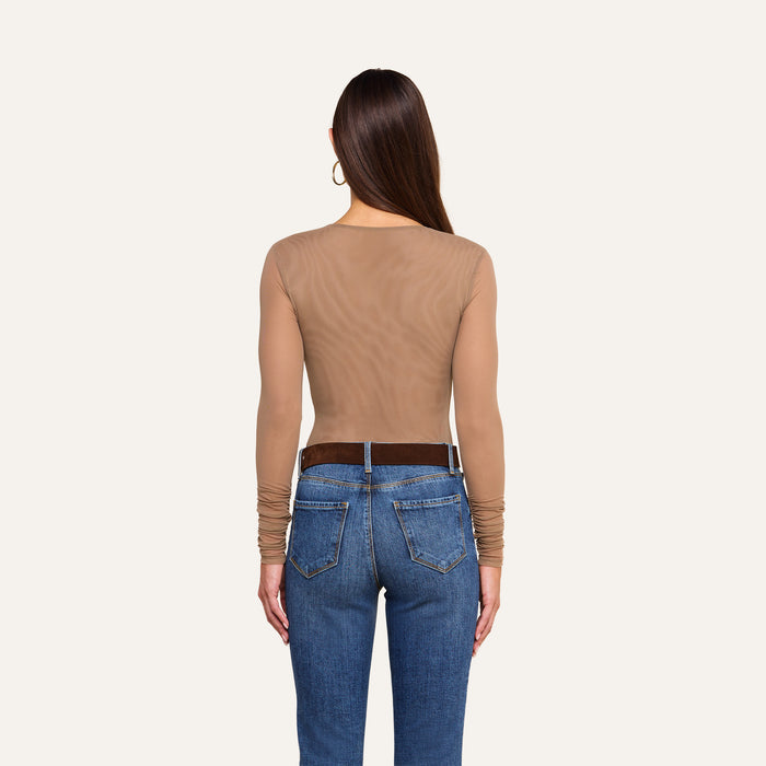 A woman stands facing away, wearing the Thalassa High-Leg Bodysuit—a sheer, long-sleeve mesh style—paired with blue jeans and a dark belt. Her long, straight hair is complemented by large hoop earrings. The background is plain and light-colored.