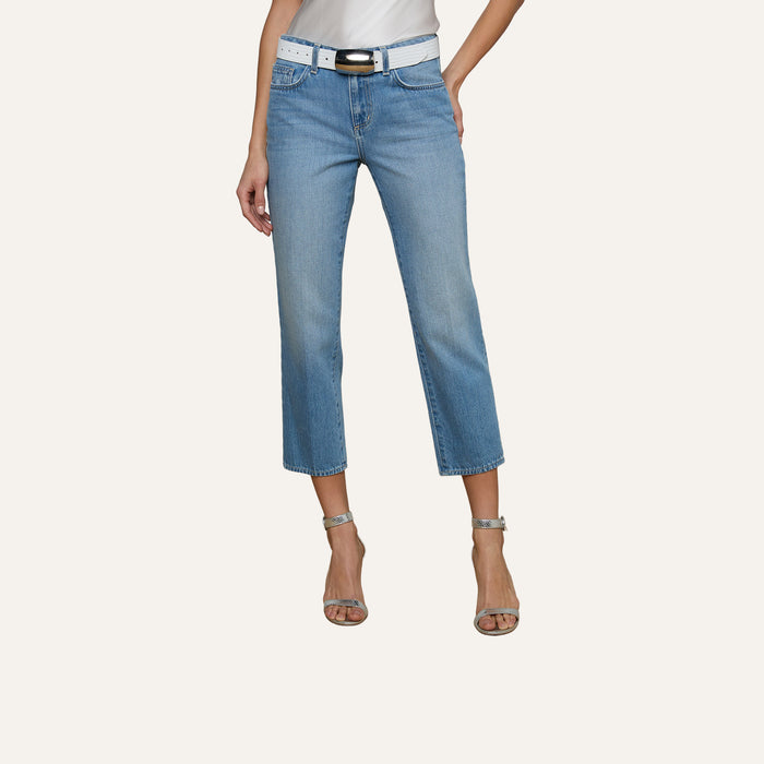 A person wearing the Thalina Cropped Stovepipe Jean in a light blue wash, paired with a white belt, a white top, and silver high-heeled sandals stands against a plain light background.