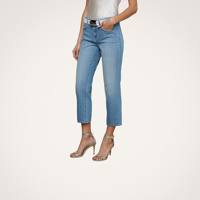 A woman stands with one hand in her pocket against a plain background, wearing the Thalina Cropped Stovepipe Jean in a light blue wash, paired with a white sleeveless top, black belt, and silver high-heeled sandals.