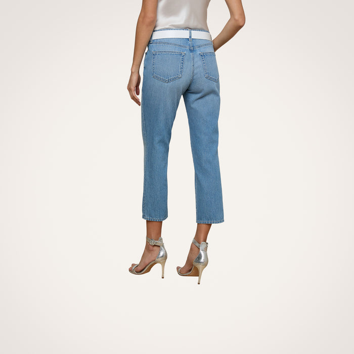 A woman stands with her back facing the camera against a plain white background, wearing the Thalina Cropped Stovepipe Jean in a light blue wash and silver high heels.