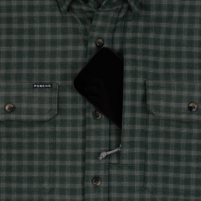 Close-up of The Acadia shirt in green plaid fabric featuring button closures and a pocket holding a black smartphone.