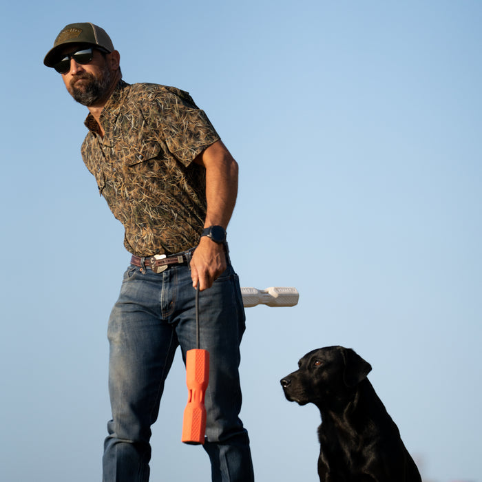 Man wearing The Layout Blind shirt outdoors holding a training dummy with a black dog sitting beside him against a clear sky