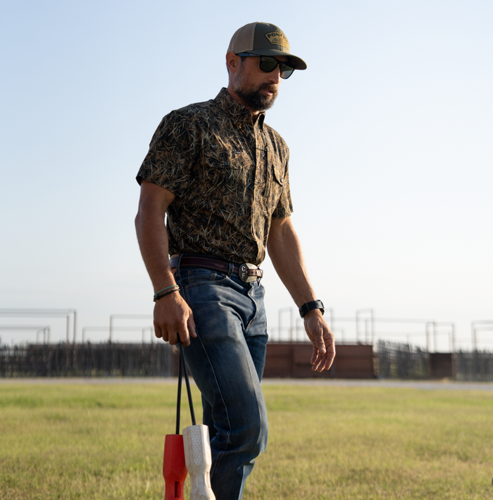 Man wearing The Layout Blind shirt outdoors paired with jeans, sunglasses, and a cap while holding two objects