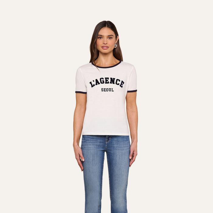 A woman stands against a plain background, wearing the Tommie Crewneck Tee—a relaxed fit white ringer tee with black trim that reads “LAGENCE SEOUL”—paired with blue jeans, looking forward with her arms relaxed by her sides.