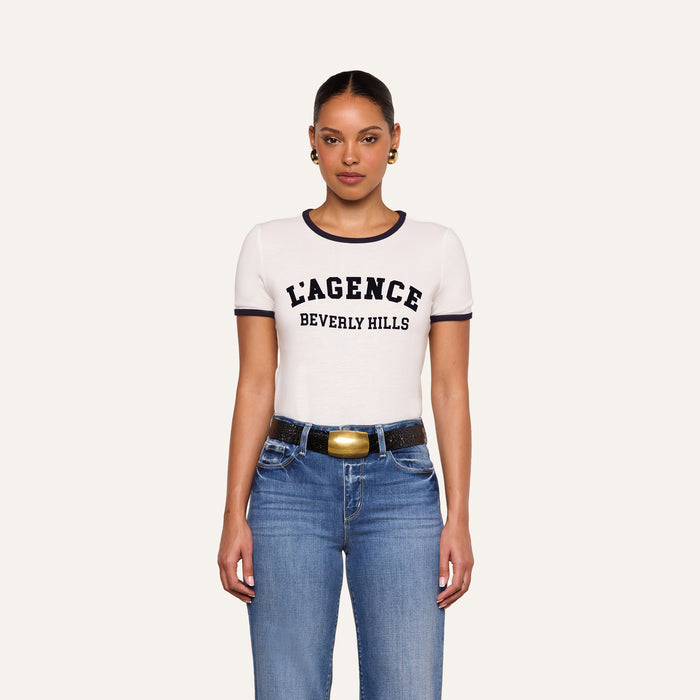 A woman stands facing forward against a plain light background, wearing the Tommie Crewneck Tee—a white Supima cotton T-shirt with “BEVERLY HILLS” in black text—paired with blue jeans, a black belt with a gold buckle, and gold earrings.