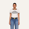 A woman stands facing forward against a plain light background, wearing the Tommie Crewneck Tee—a white Supima cotton T-shirt with “BEVERLY HILLS” in black text—paired with blue jeans, a black belt with a gold buckle, and gold earrings.