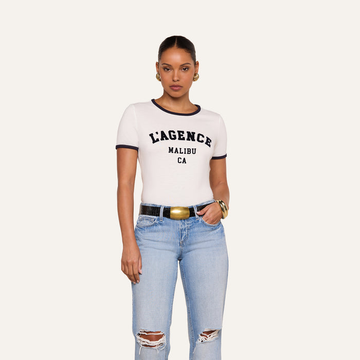 A woman stands against a plain background, looking directly at the camera, wearing hoop earrings, gold bangles, ripped light blue jeans with a black belt, and a white Tommie Crewneck Tee featuring L’AGENCE MALIBU CA text.