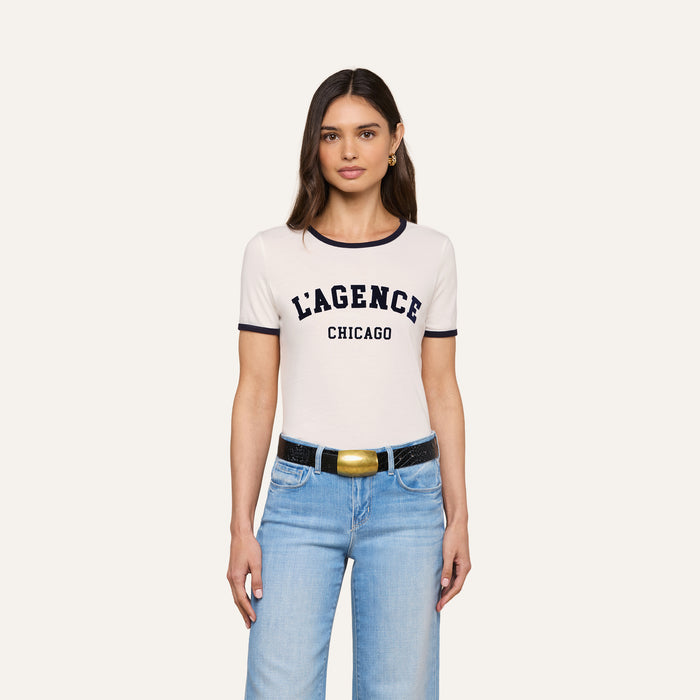Woman with long brown hair wearing the Tommie Crewneck Tee in white, paired with light blue jeans and a black belt with a gold buckle, standing against a plain light background.