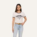 A woman wearing the Tommie Crewneck Tee—a relaxed fit white ringer tee made from Supima cotton featuring LAGENCE NEWPORT CA—paired with light blue jeans, a tan belt with a gold buckle, and gold hoop earrings, stands against a plain background.