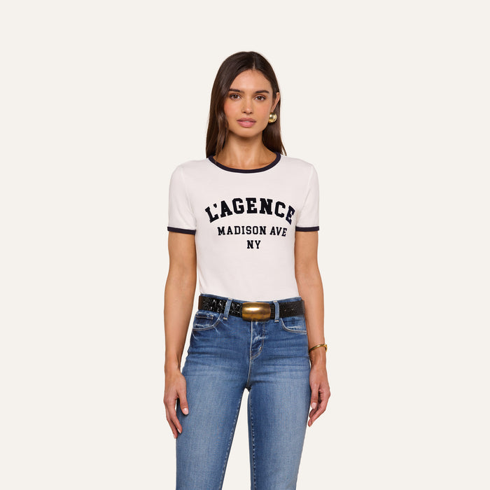 A woman with long brown hair wears a relaxed fit white Tommie Crewneck Tee made from Supima cotton with LAGENCE MADISON AVE NY printed on it, blue jeans, a black belt, and gold hoop earrings, standing against a plain, light background.