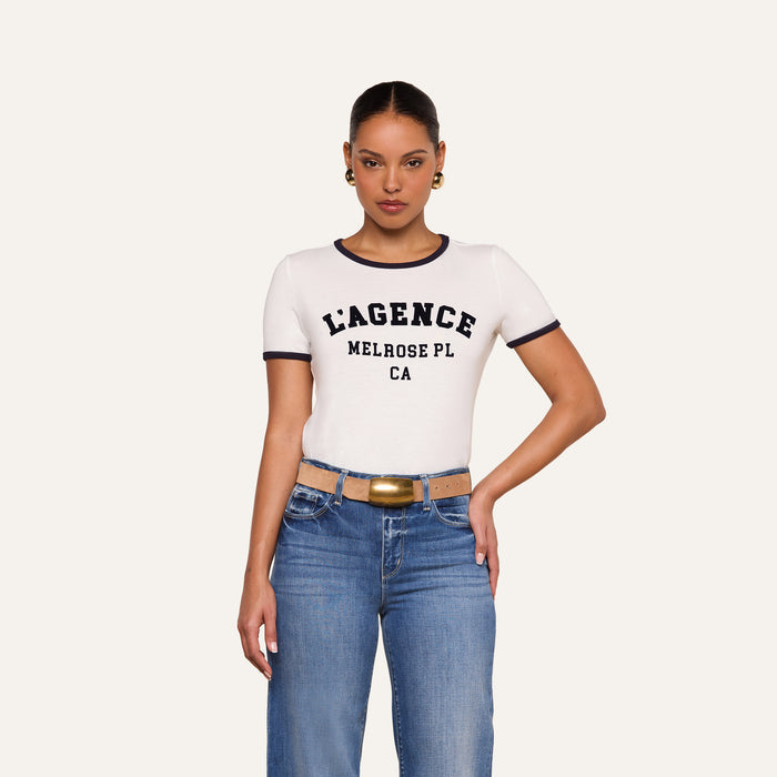 Woman in a relaxed fit Tommie Crewneck Tee with LAGENCE MELROSE PL CA text, blue jeans, a gold-buckled belt, and hoop earrings poses against a plain light background.