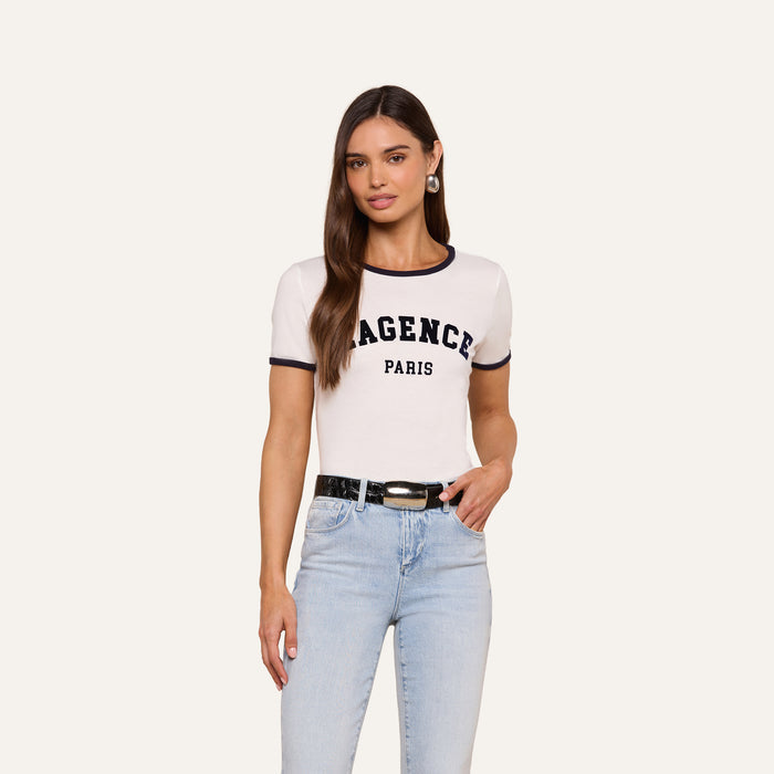 A woman with long brown hair wears the Tommie Crewneck Tee— a white, short-sleeve Paris top made from soft Supima cotton that says AGENCE PARIS—paired with light blue jeans, a black belt, and hoop earrings, standing against a plain light background.
