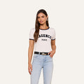 A woman with long brown hair wears the Tommie Crewneck Tee— a white, short-sleeve Paris top made from soft Supima cotton that says AGENCE PARIS—paired with light blue jeans, a black belt, and hoop earrings, standing against a plain light background.