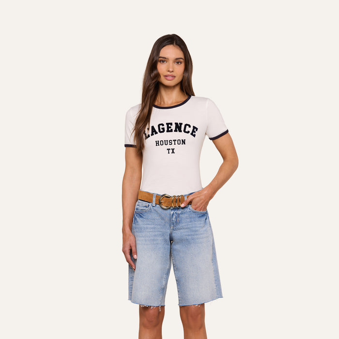A woman is wearing a relaxed fit white Tommie Crewneck Tee made of Supima cotton featuring the “AGENCE HOUSTON TX” text, paired with blue denim knee-length shorts, standing with one hand in her pocket against a plain background.