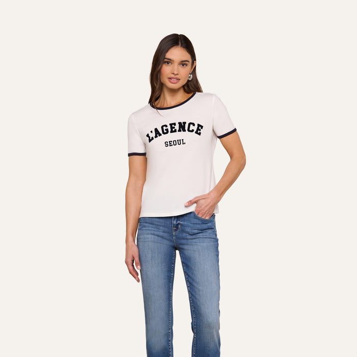 A woman stands against a plain background, wearing the Tommie Crewneck Tee—a white ringer style with “LAGENCE SEOUL” printed on the front—paired with blue jeans. She has one hand in her pocket and a relaxed expression.