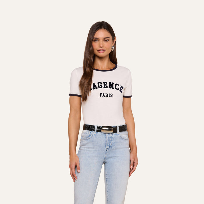 A woman with long brown hair wears a white Tommie Crewneck Tee crafted from Supima cotton, styled with light blue jeans and a black belt, standing against a plain light background.