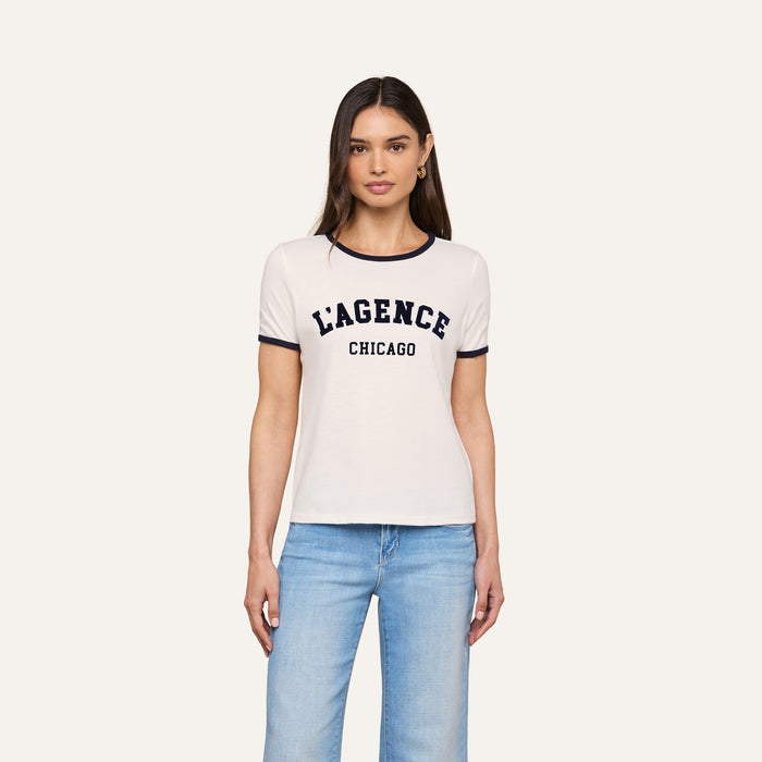 A woman stands against a plain background, wearing the Tommie Crewneck Tee—a relaxed fit white ringer tee crafted from Supima cotton with LAGENCE CHICAGO printed on it—paired with light blue jeans.