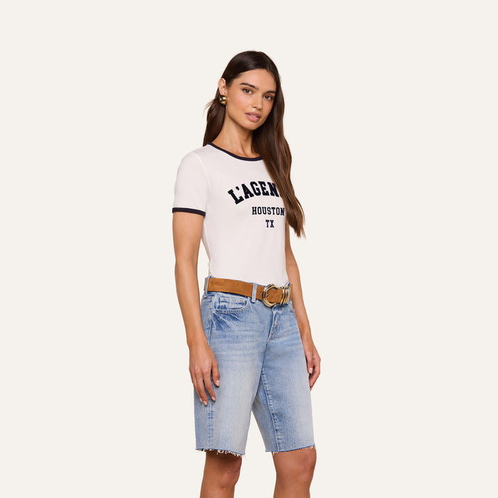 A woman with long brown hair wears a white Tommie Crewneck Tee crafted from Supima cotton, denim Bermuda shorts, a brown belt, and earrings, standing against a plain light background.