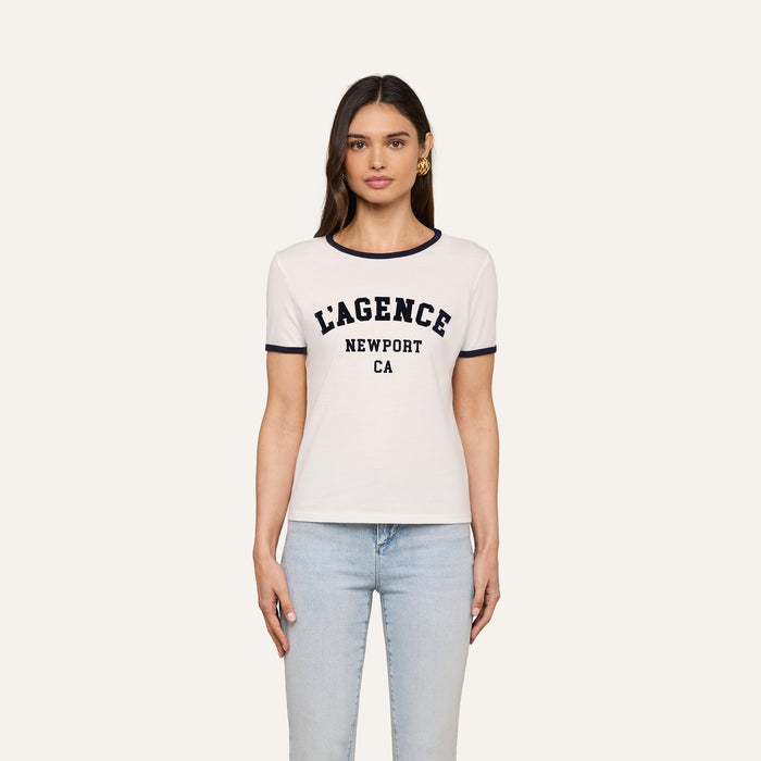 A woman is wearing the Tommie Crewneck Tee—a white short-sleeve ringer shirt with black trim and “LAGENCE NEWPORT CA” printed on the front, crafted from soft Supima cotton—paired with light blue jeans and standing against a plain white background.