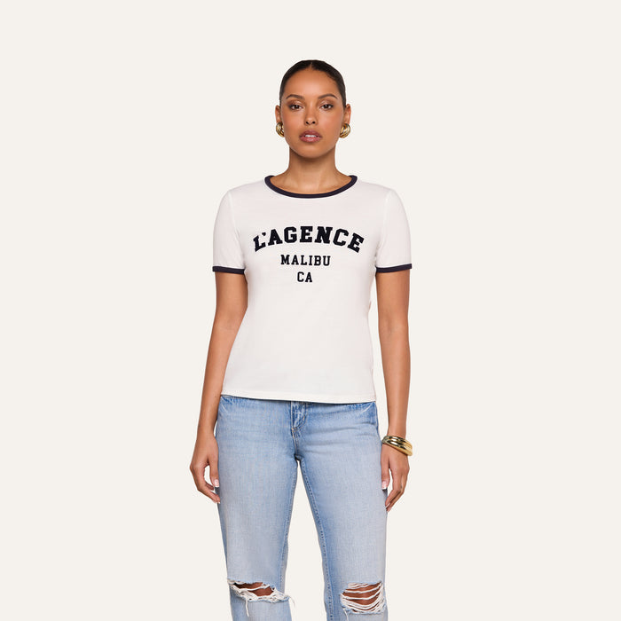 A woman wearing a white Tommie Crewneck Tee with the L’AGENCE logo MALIBU CA text and blue jeans with ripped knees stands facing forward against a plain background.