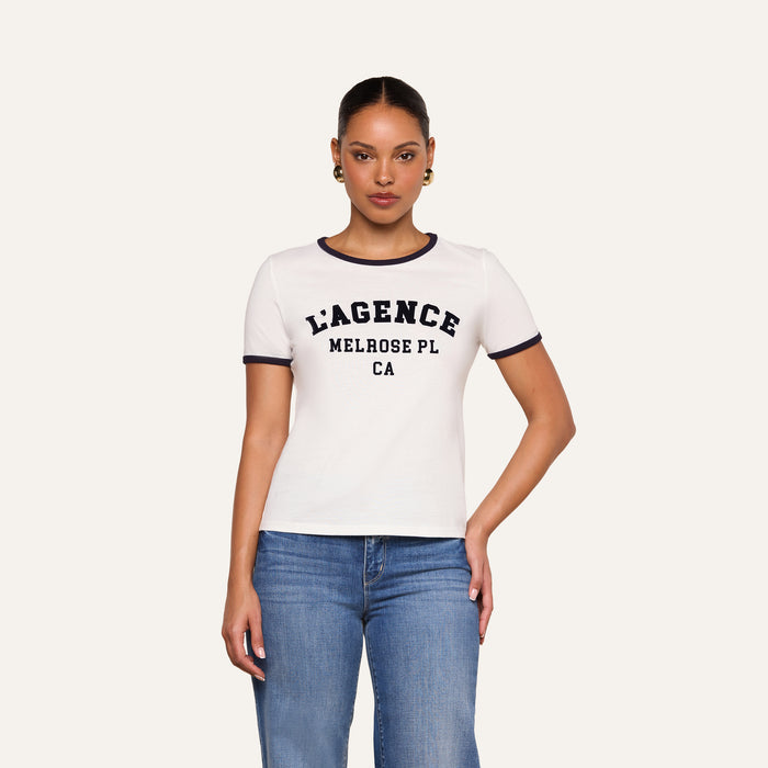 A woman wearing the Tommie Crewneck Tee—a relaxed-fit ringer style crafted from Supima cotton with dark trim and “LAGENCE MELROSE PL CA” text—paired with blue jeans, stands against a plain light background.
