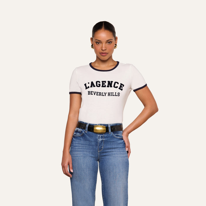 A woman wearing the Tommie Crewneck Tee, a relaxed fit white Supima cotton T-shirt with “Beverly Hills” text, paired with blue jeans, stands against a plain light background with one hand resting on her hip.