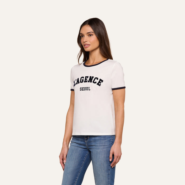 A woman wearing a white Tommie Crewneck Tee made from Supima cotton, featuring “LAGENCE SEOUL” printed on the front, and blue jeans stands against a plain background, gazing slightly to her right. The relaxed fit of the tee enhances her casual, effortless style.