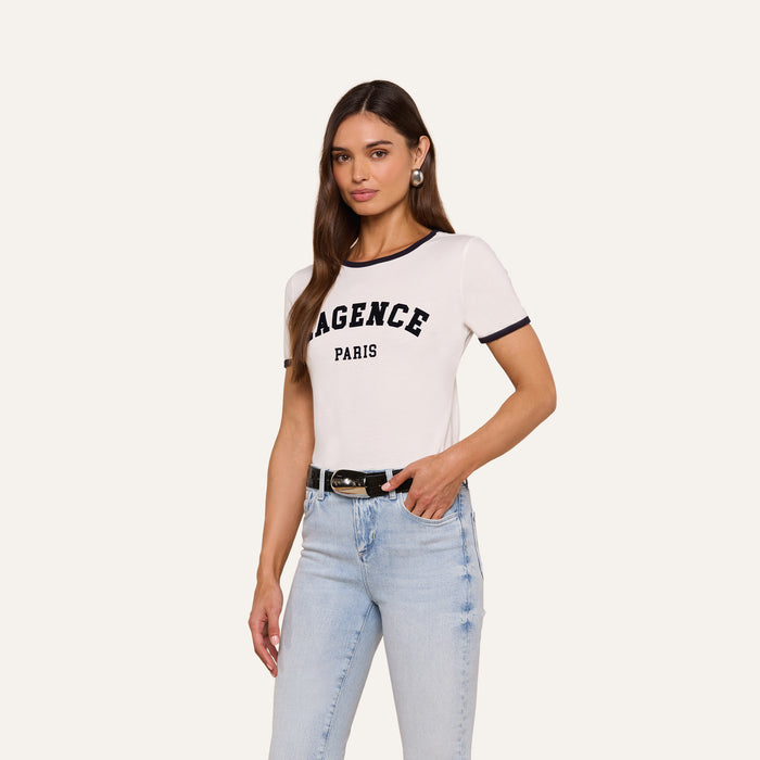 Woman wearing the Tommie Crewneck Tee in white with LAGENCE PARIS text, light blue jeans, black belt, and hoop earrings, standing against a plain light background in a relaxed fit Supima cotton style.