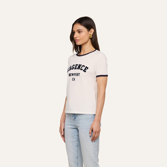 A woman wearing the Tommie Crewneck Tee, a white ringer t-shirt with “LAGENCE NEWPORT CA” text, paired with relaxed fit light blue jeans and gold earrings, stands against a plain light background facing slightly to the side.