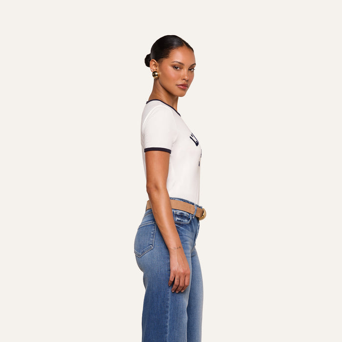 A woman stands in profile against a plain background, wearing the Tommie Crewneck Tee—a relaxed fit ringer style crafted from Supima cotton with black trim—paired with blue jeans, a brown belt, and gold earrings. Her hair is styled in a low, sleek bun.