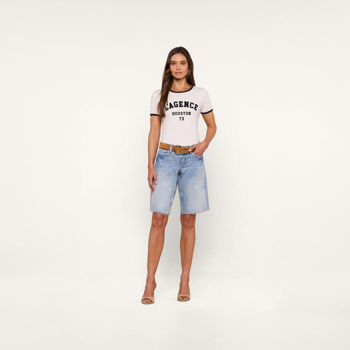 A woman stands facing forward, wearing a relaxed fit Tommie Crewneck Tee—a white T-shirt with black trim and AGENCE HOUSTON TX text—paired with light blue knee-length denim shorts, a brown belt, and beige heels. The background is plain white.