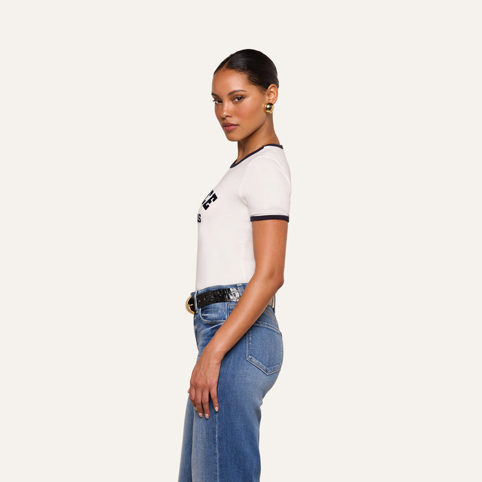 Woman standing in profile, wearing a relaxed fit white Tommie Crewneck Tee with black trim, blue jeans, a black belt with a gold buckle, and gold earrings, against a plain light background.