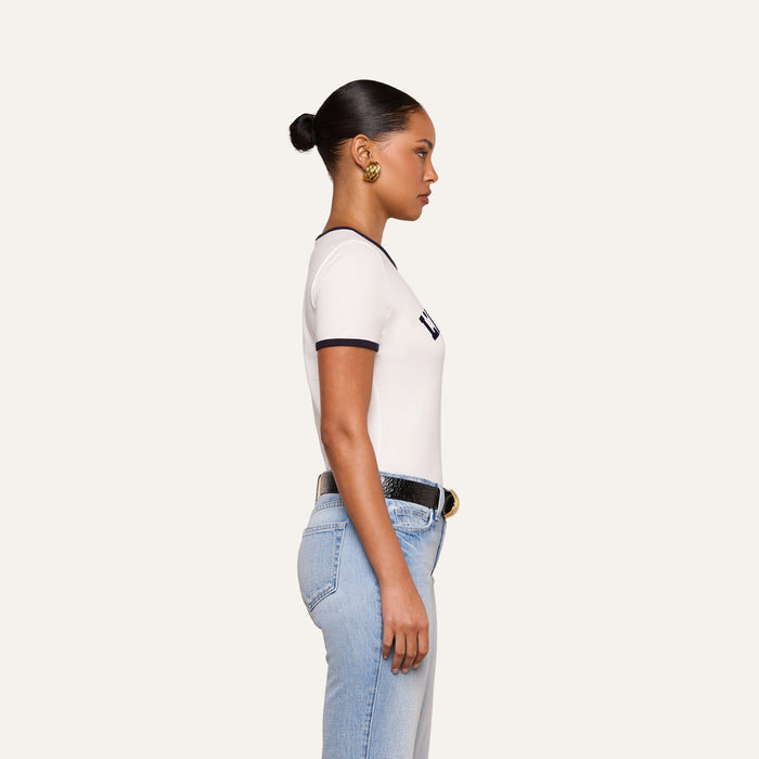 A woman stands in profile against a plain white background, wearing the Tommie Crewneck Tee—a white ringer tee with black trim—paired with light blue jeans, a black belt, gold earrings, and her hair styled in a low bun.