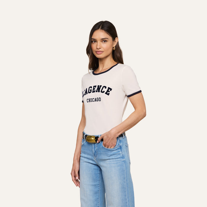 A woman with long brown hair wears the Tommie Crewneck Tee—a relaxed fit white ringer shirt with “AGENCE CHICAGO” on the front—paired with light blue jeans and a black belt, posing with one hand in her pocket against a plain background.