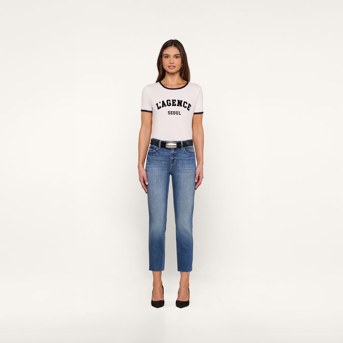 A woman stands facing forward, wearing a white Tommie Crewneck Tee in Supima cotton with a relaxed fit, blue jeans, a black belt, and black high heels against a plain white background.