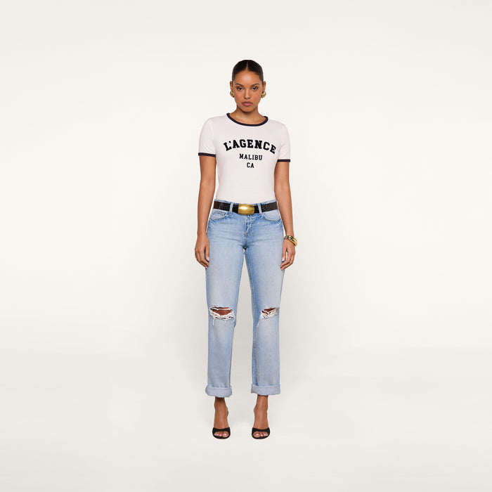 A woman stands against a plain background, wearing a white Tommie Crewneck Tee made from Supima cotton featuring the L’AGENCE logo, light blue ripped jeans, black heeled sandals, a black belt, hoop earrings, and gold bracelets.