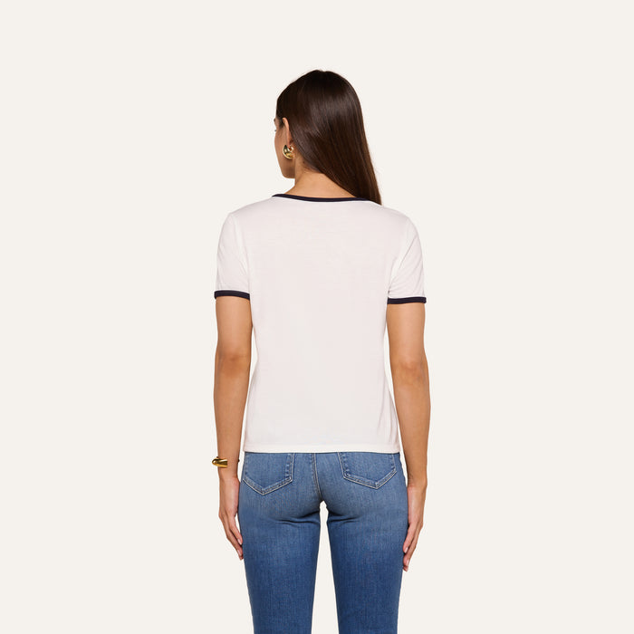 A woman with long brown hair, wearing a relaxed fit Tommie Crewneck Tee made from Supima cotton with black trim and blue jeans, stands facing away from the camera against a plain background.